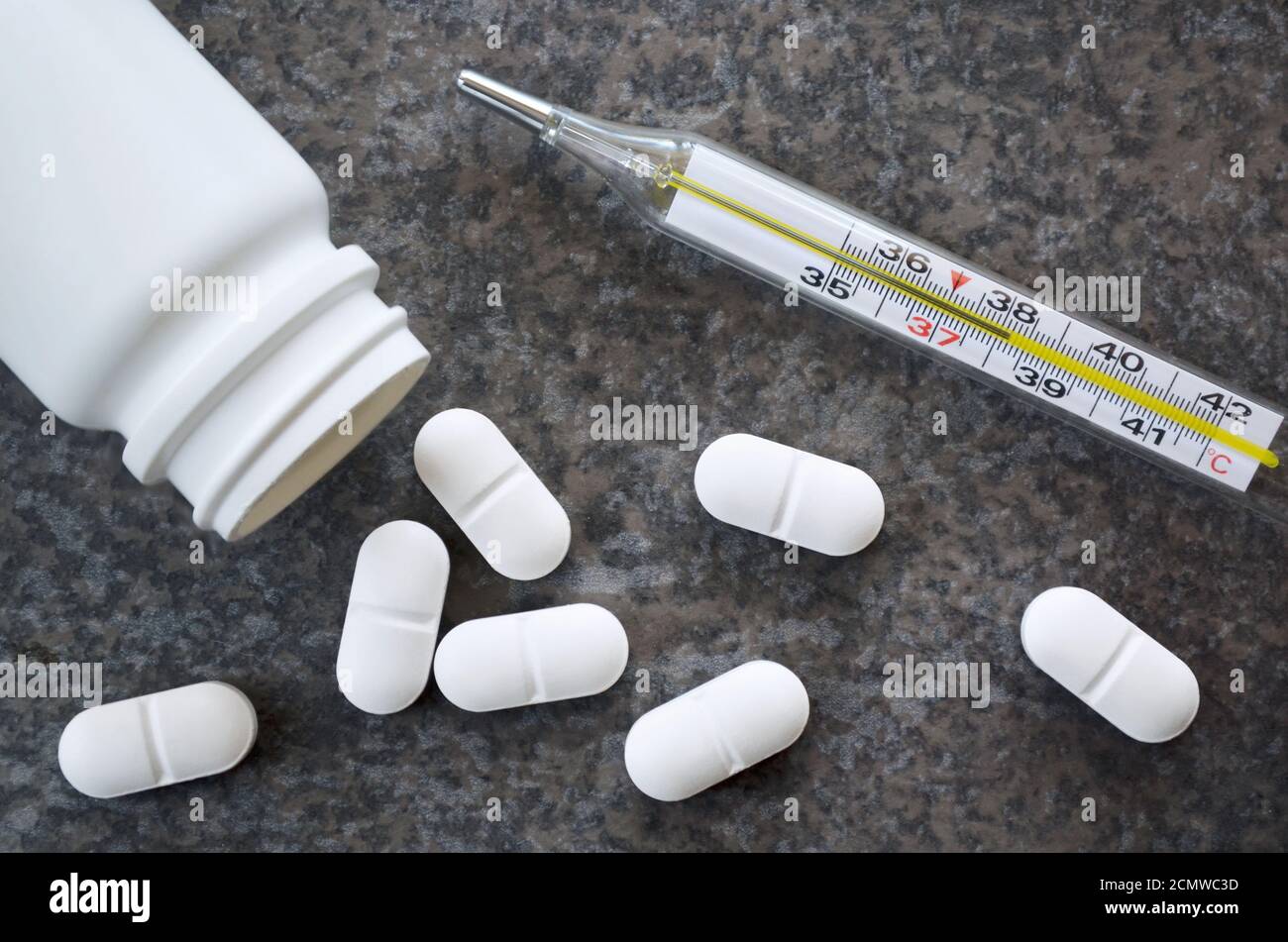 White pills and a medical mercury-in-glass thermometer indicating a critical temperature of 38.2 degrees Celsius on a dark gray table surface. Stock Photo