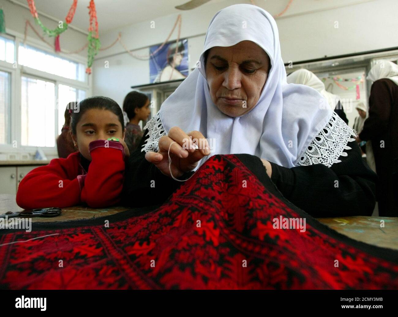 A Palestinian woman is watched by a young girl as she embroiders in a women programme center in Gaza City January 5, 2003, where women are taught hairdressing, stitching and embroidery and IT skills. Palestinian women have been exposed to increased domestic violence since the beginning of the intifada, or Palestinian uprising, 29 months ago, according to the Palestinian Center for Public Opinion (PCPO), which was commissioned by a Palestinian women's organization to conduct a poll. The poll showed that 86 percent of respondents said violence against women had significantly or somewhat increase Stock Photo