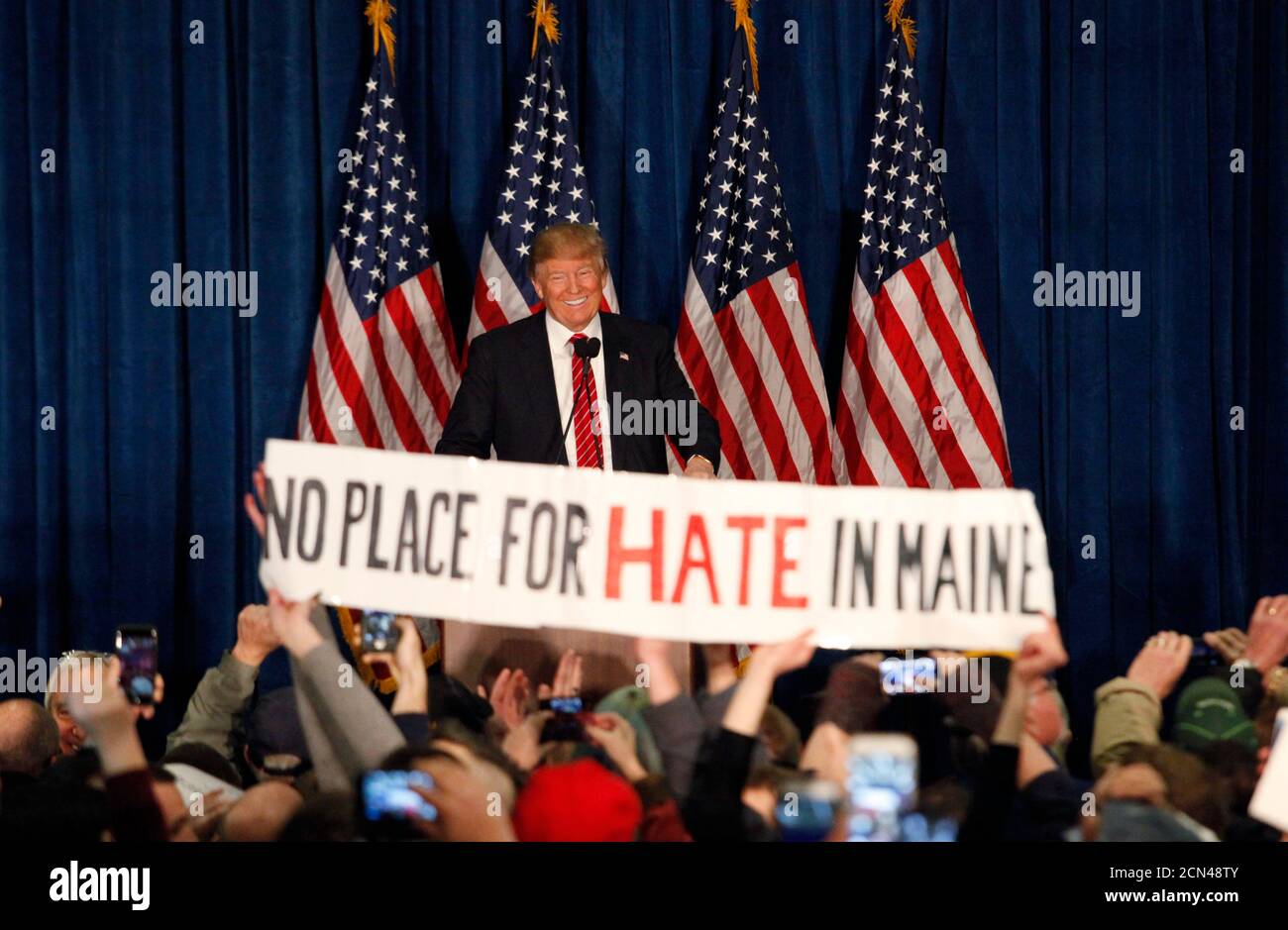 Republican U.S. presidential candidate Donald Trump smiles as protestors hold up a sign reading 'No Place for Hate in Maine' during a campaign rally in Portland, Maine March 3, 2016.  REUTERS/Joel Page Stock Photo