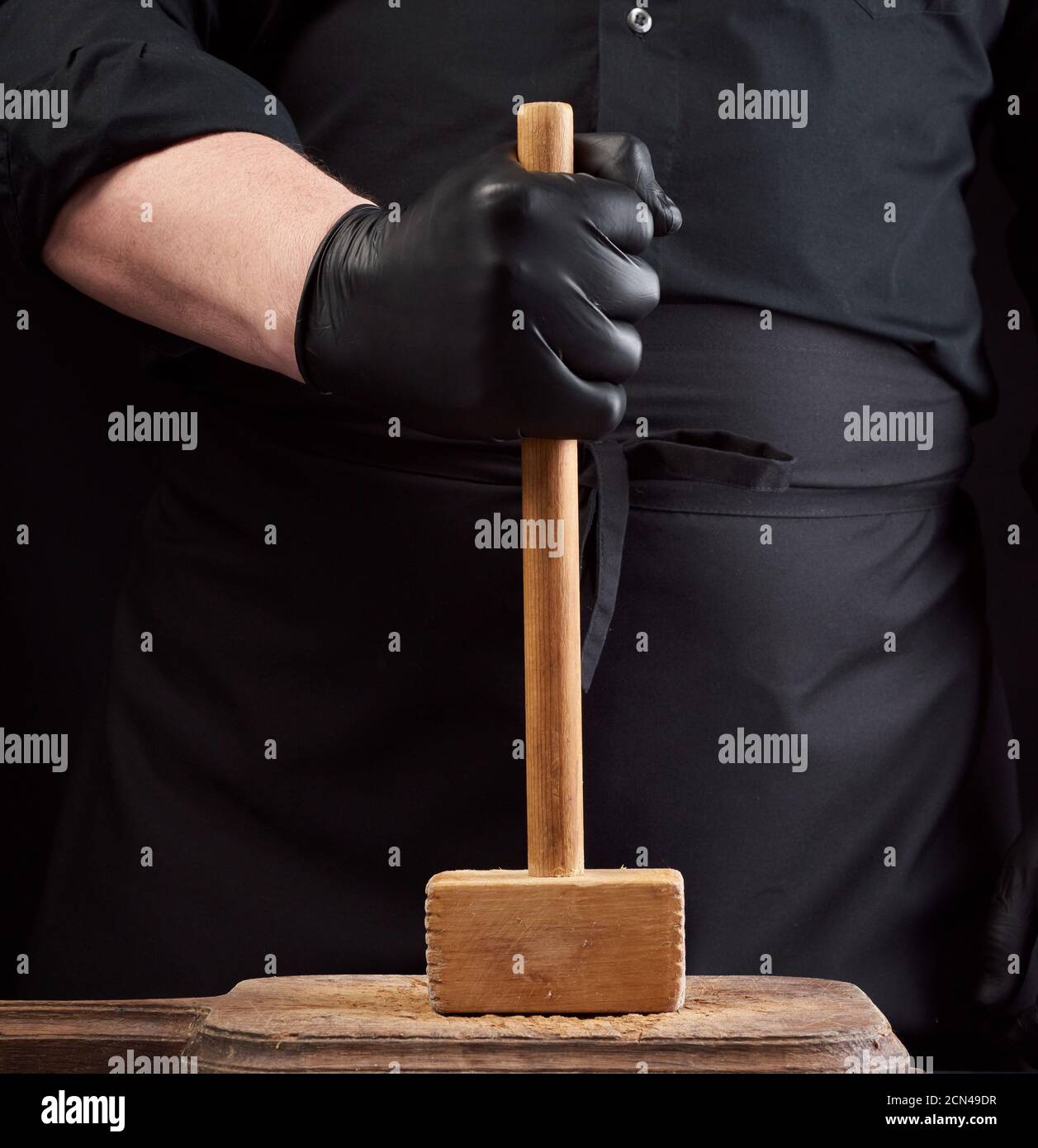 cook in black uniform holds a wooden hammer for beating meat on a ...