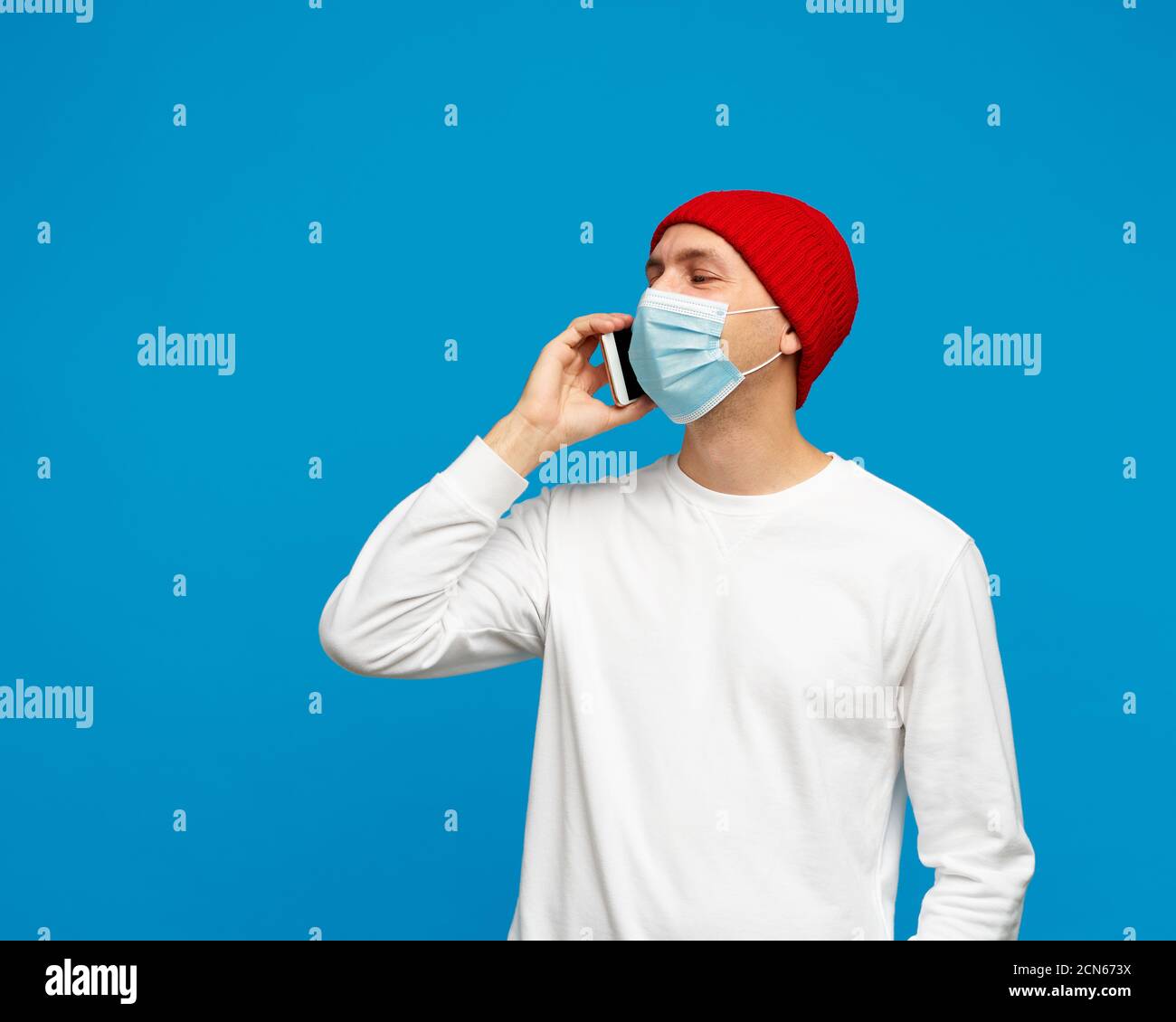 Portrait of man with medical protective face mask, speaking to mobile phone. Male isolated on bright blue colored background. White jumper and red hat. Stock Photo