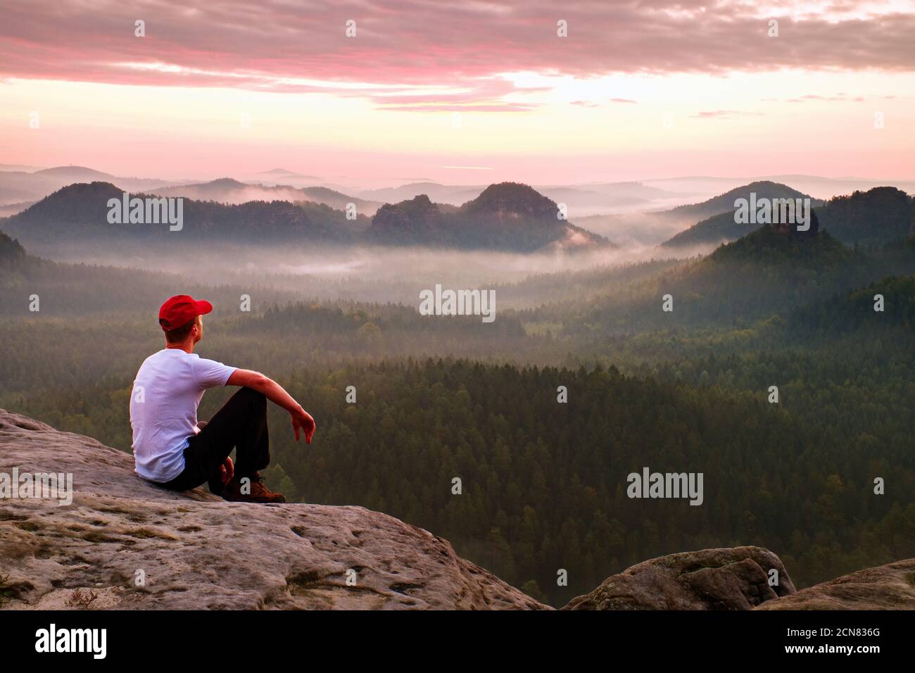 Adult sportsman in white shirt, dark trousars and red cap. Ginger short hair man sit on sharp cliff above valley in rocky mounta Stock Photo