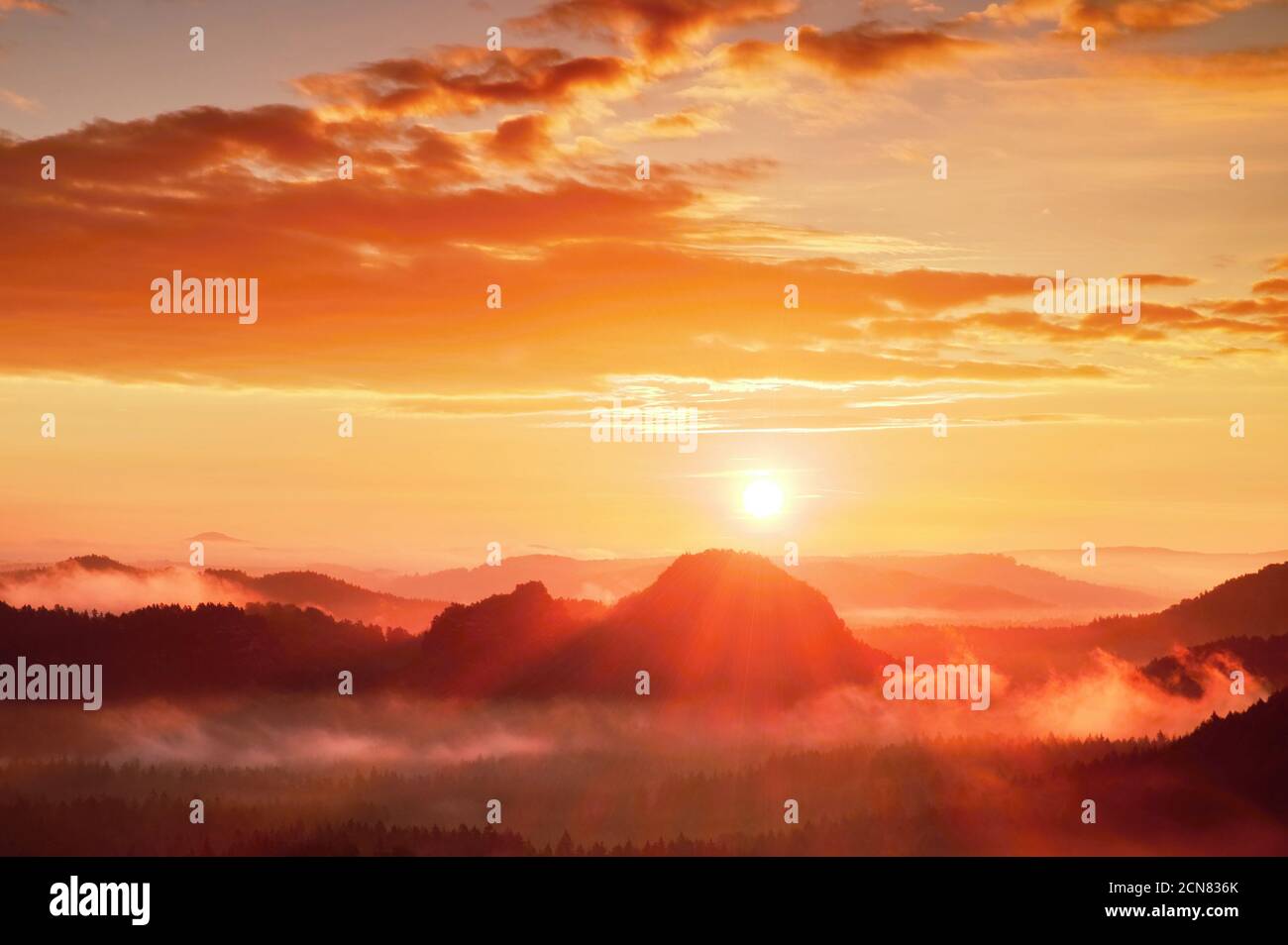 Red misty daybreak. Foggy autumn morning in a beautiful hills. Peaks of hills are sticking out from rich colorful clouds. Stock Photo