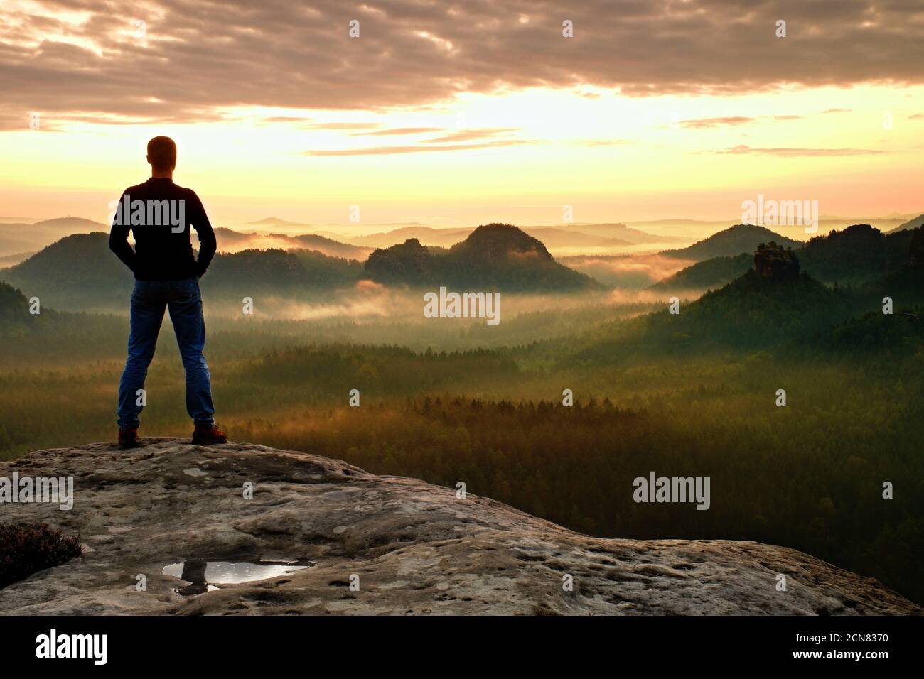 Alone man is standing on peak of sharp cliff in rock empires park and watching over the misty and foggy morning valley to Sun. B Stock Photo