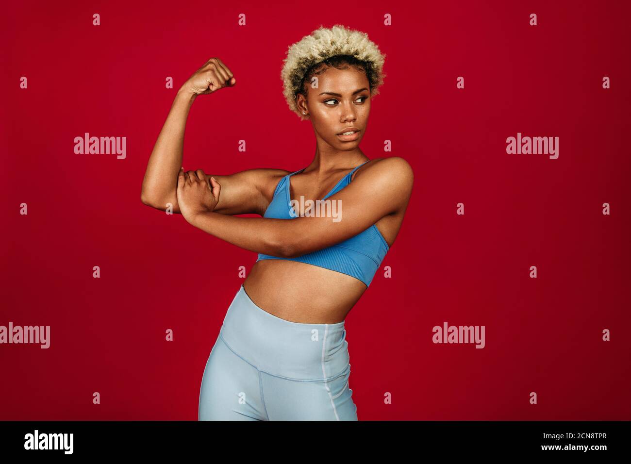 African american fitness woman standing against red background showing biceps. Portrait of woman in fitness wear doing workout. Stock Photo