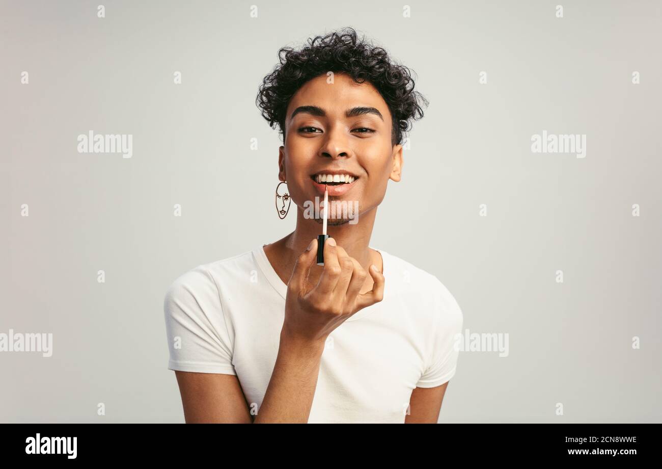 Young androgynous man applying makeup on his lips. Gay man putting on lip gloss with applicator against white background. Stock Photo