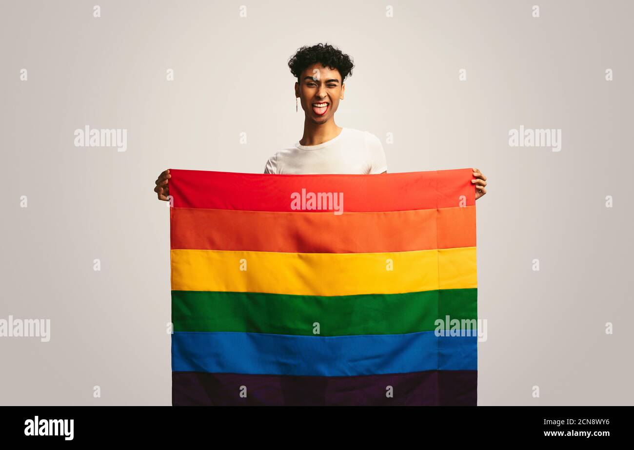 Man sticking out tongue and winking holding pride flag. Gay man with lgbt flag making funny face on white background. Stock Photo
