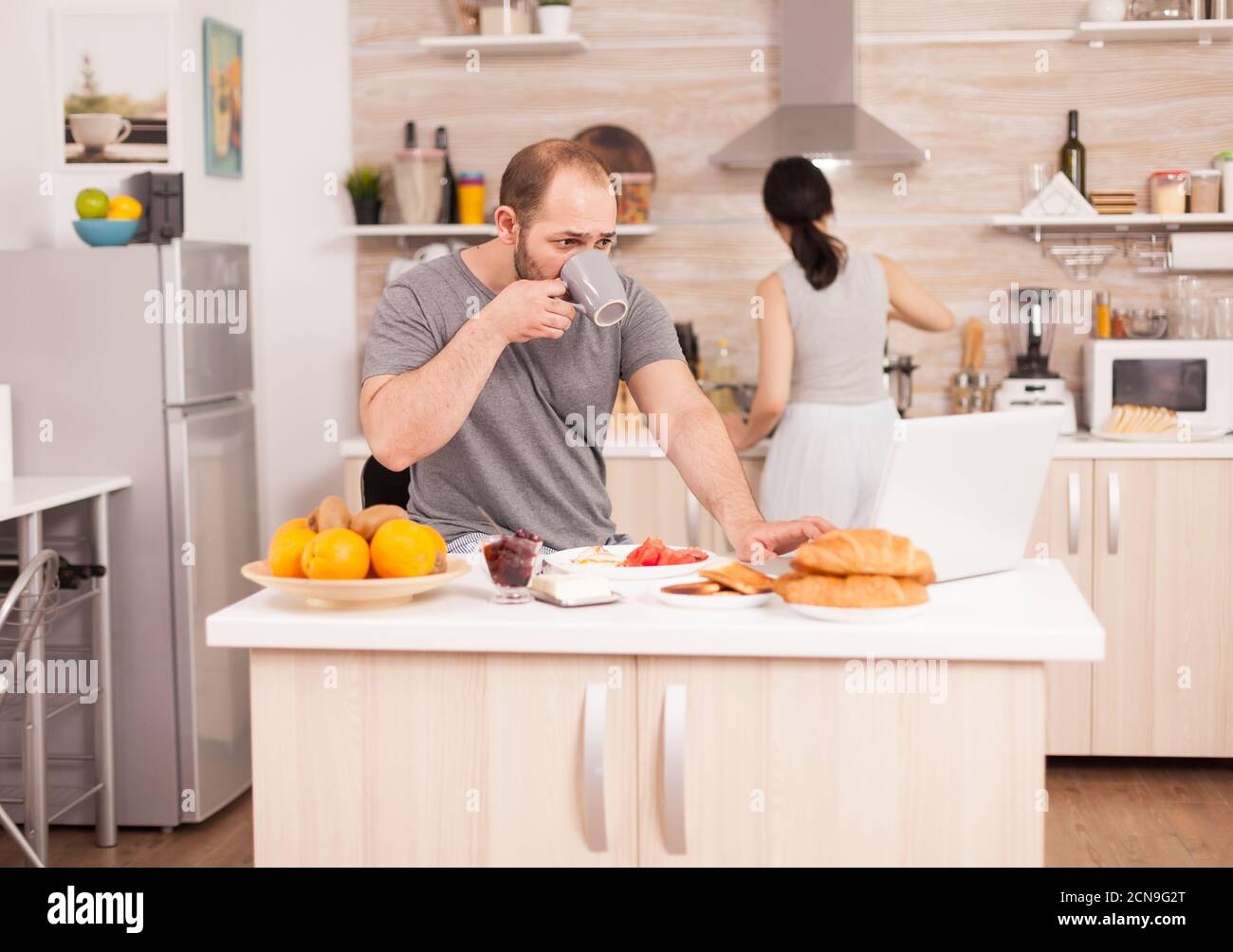 Freelancer drinking coffee and working from home while his wife prepares the breakfast. Freelancer work online via internet using modern digital technology Stock Photo