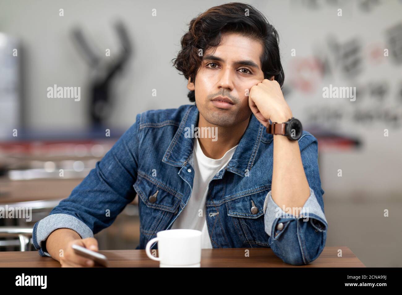 Portrait of a young handsome Indian man with serious look, lost in thought, sitting in an office cafeteria, coffee shop, casual work environment. Stock Photo