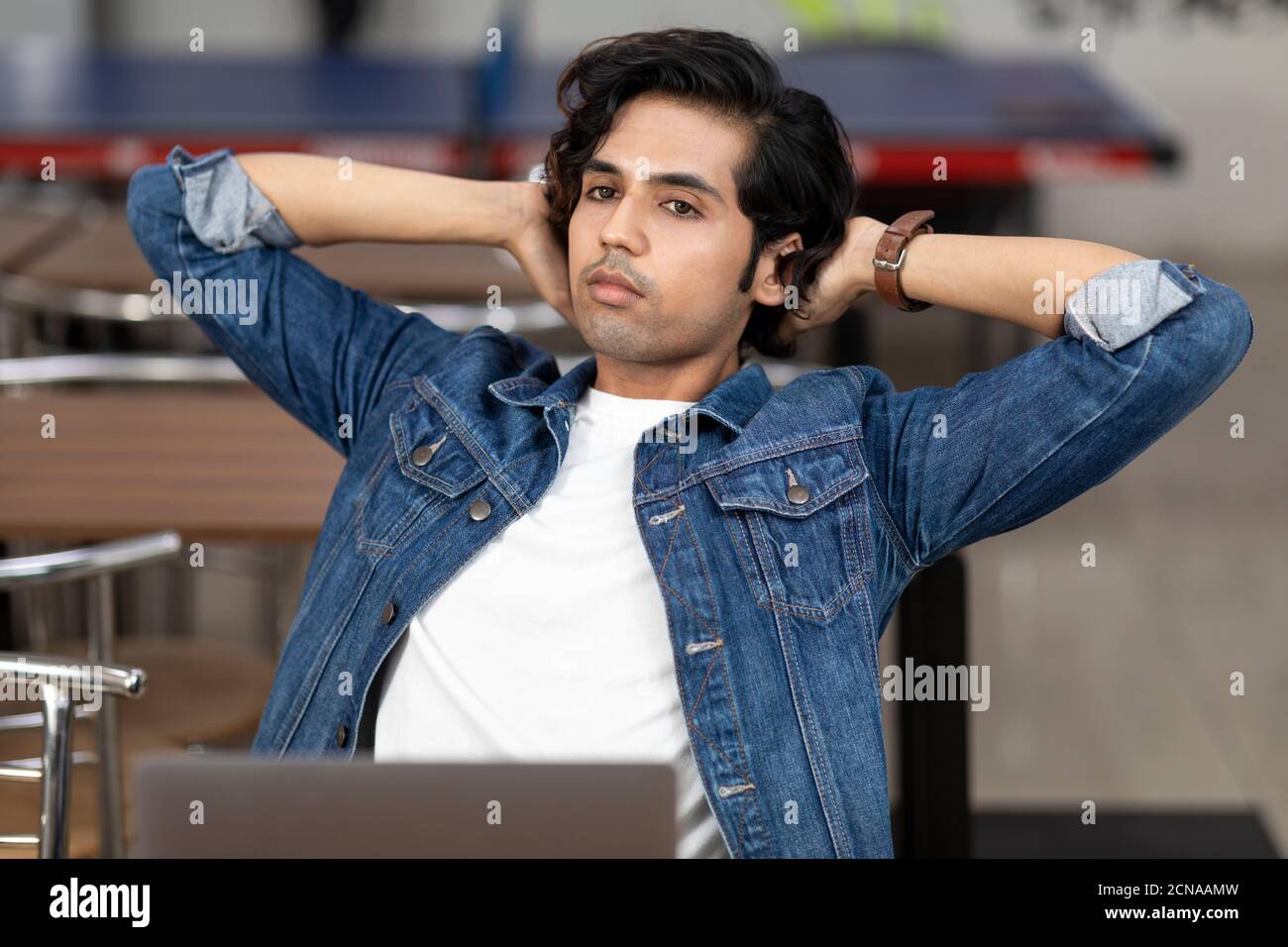 Portrait of a tired Indian man working on his laptop, sitting in an office cafeteria, coffee shop, casual work  head resting back, lost in thought. Stock Photo