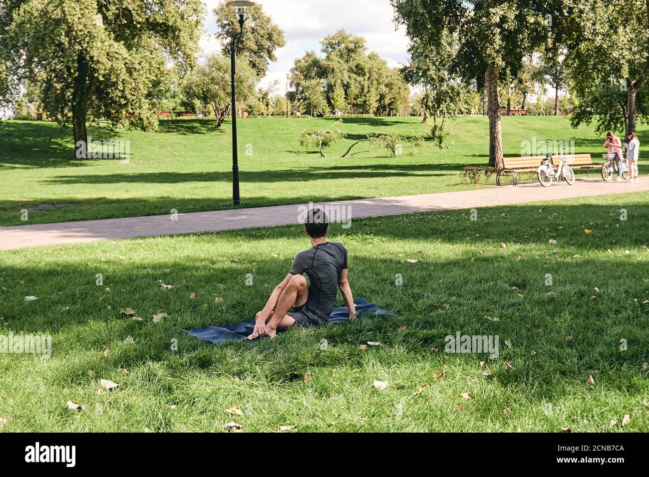 Young athletic man in sportswear doing yoga in the park. Practice half spinal twist asana outdoors. People exercising stretching on green grass with Stock Photo