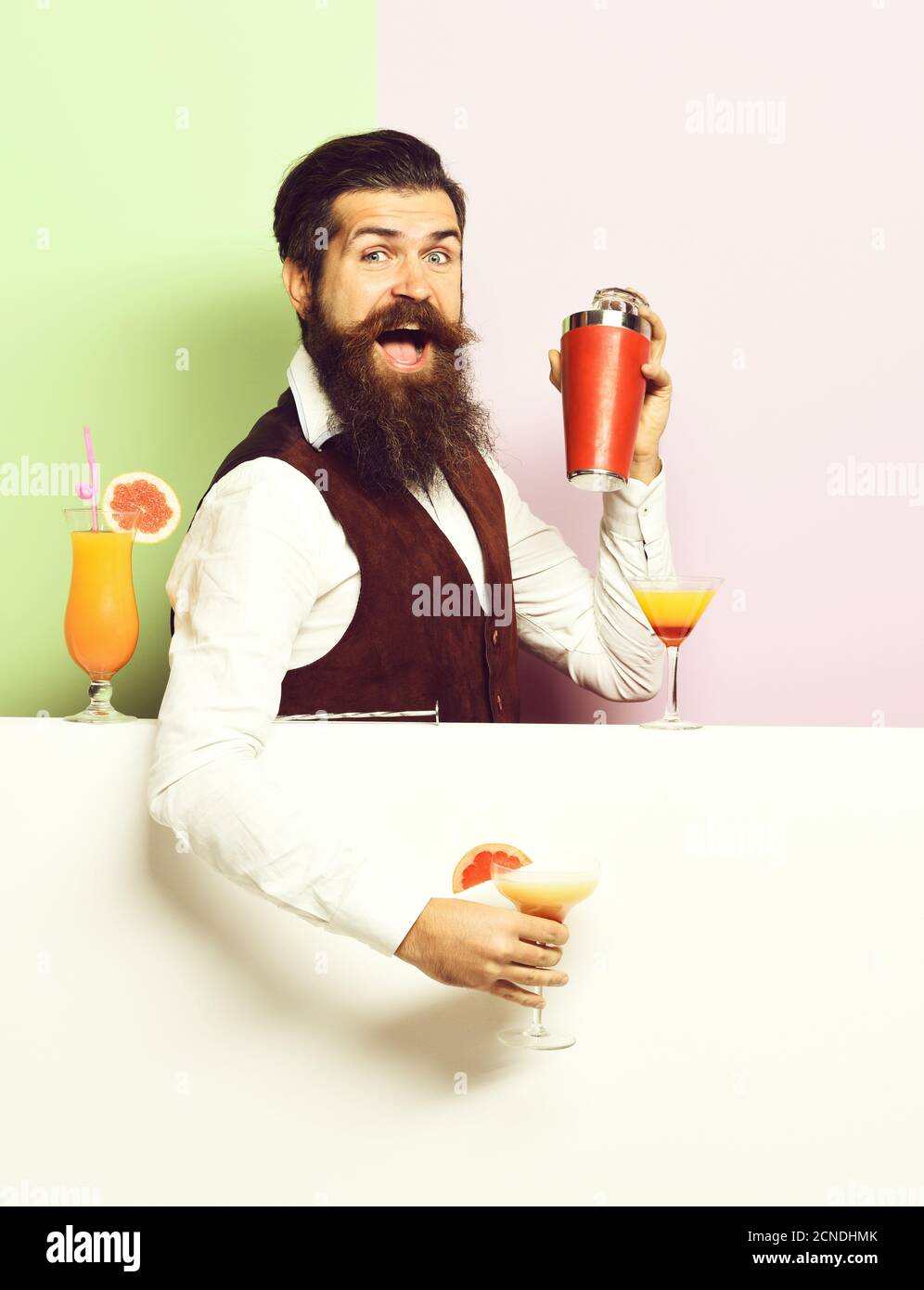 handsome bearded barman with long beard and mustache has stylish hair on funny face holding shaker and made alcoholic cocktail in vintage suede leather waistcoat, on purple green studio background Stock Photo