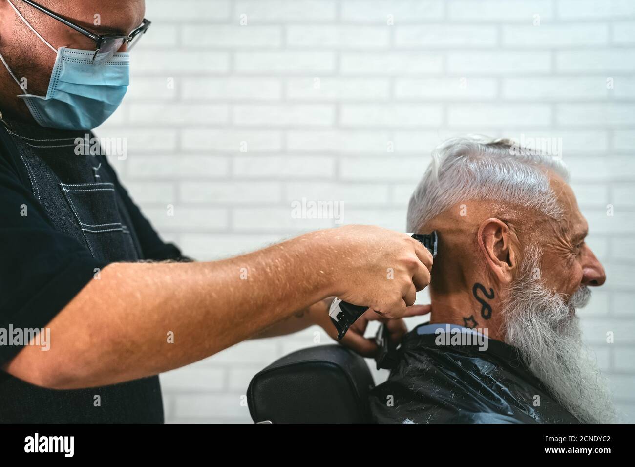 Male hairdresser cutting hair to hipster senior client while wearing face surgical mask - Young hairstylist working in barbershop during corona virus Stock Photo