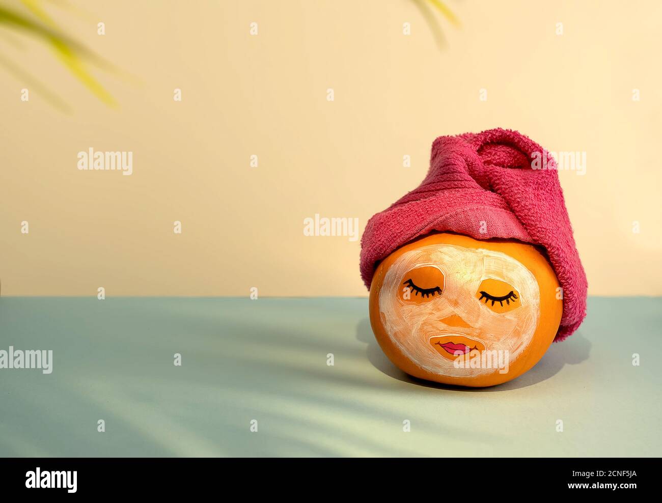 Pumpkin with painted face and facial mask, towel and flowers on pastel yellow and blue background with shadow from palm leaves. Spa concept, Helloween, Copy space. Stock Photo