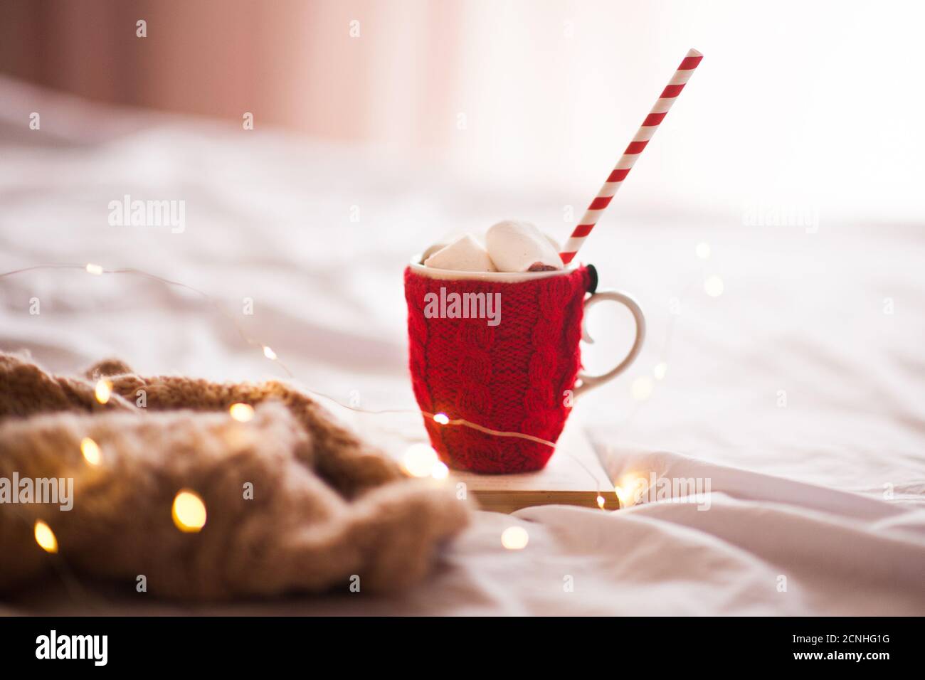 Tasty cup of coffee with red knitted textile with marshmallow and water pipe staying on open book in bed close up. Winter holiday season. Xmas. Good m Stock Photo