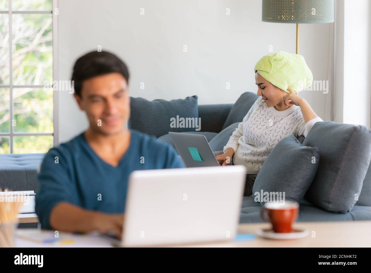Asian businesswoman with towel binding on head sitting and using notebook computer on sofa, blur Caucasian businessman using laptop  working at desk. Stock Photo