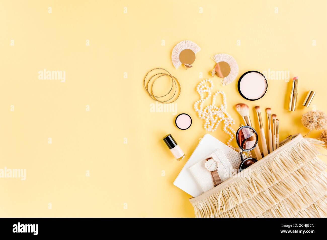 Woman straw bag with travel accessories, makeup brushes, cosmetics, golden accessories on yellow background.  Stock Photo