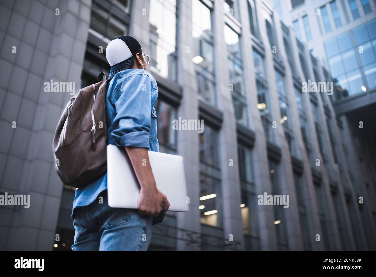 Cheerful young man with backpack enjoying walk the city with laptop and snap back. Stock Photo