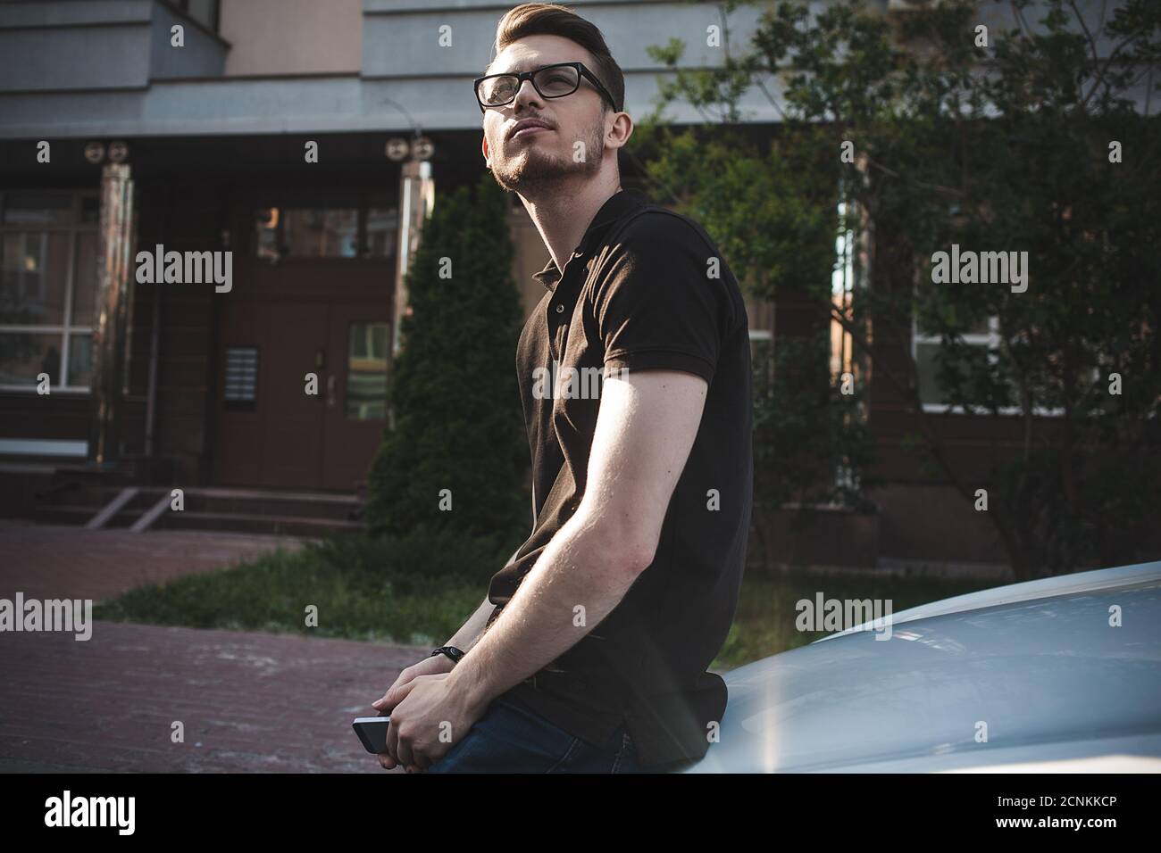 Young man leaning on his car, using a smartphone, dressed casually. Stock Photo