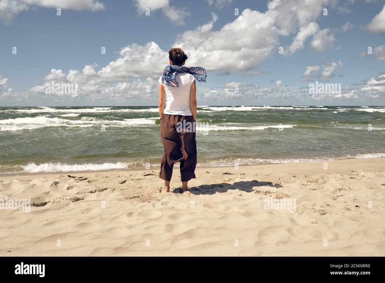 Lonely middle aged stylish woman from behind standing on the beach and looking at sea on windy summer day Stock Photo
