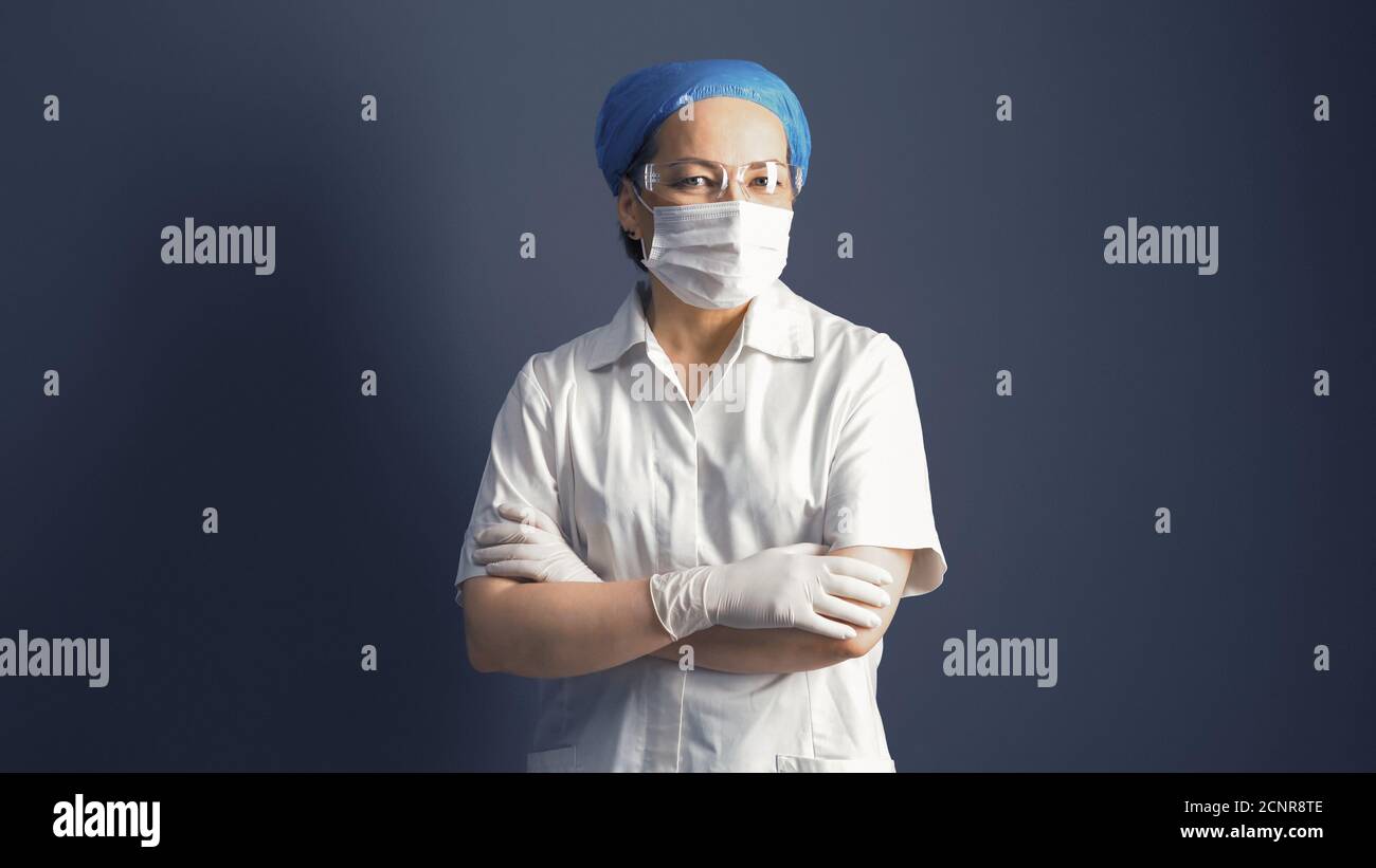 Female doctor looks at camera crossed arms. Female medic in mask and white uniform, portrait on gray background Stock Photo