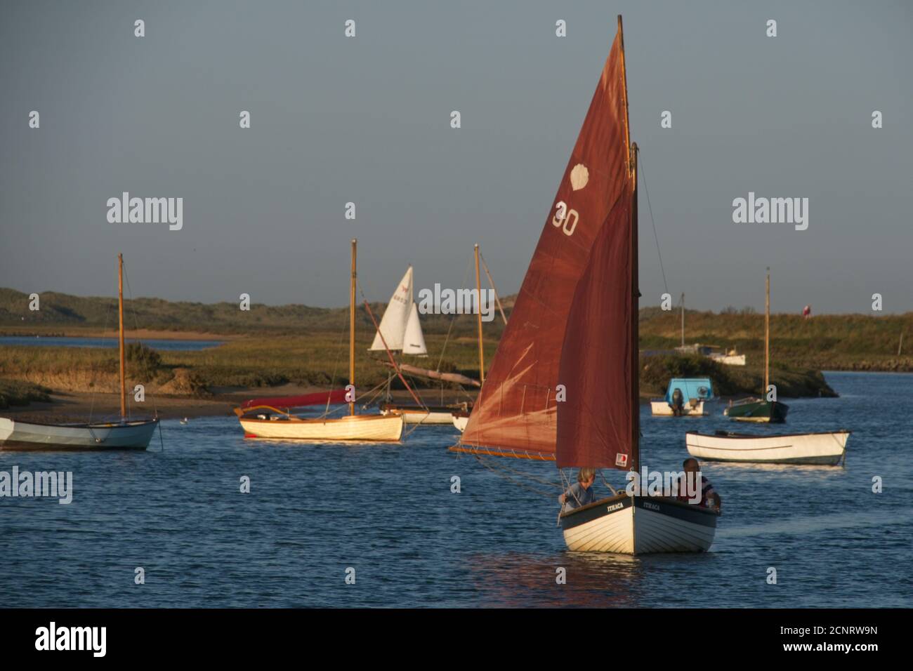 Traditional sail boat under sail in estuary among boats at moorings.  Plain grey sky. Boats moored in background. Landscape format. Stock Photo