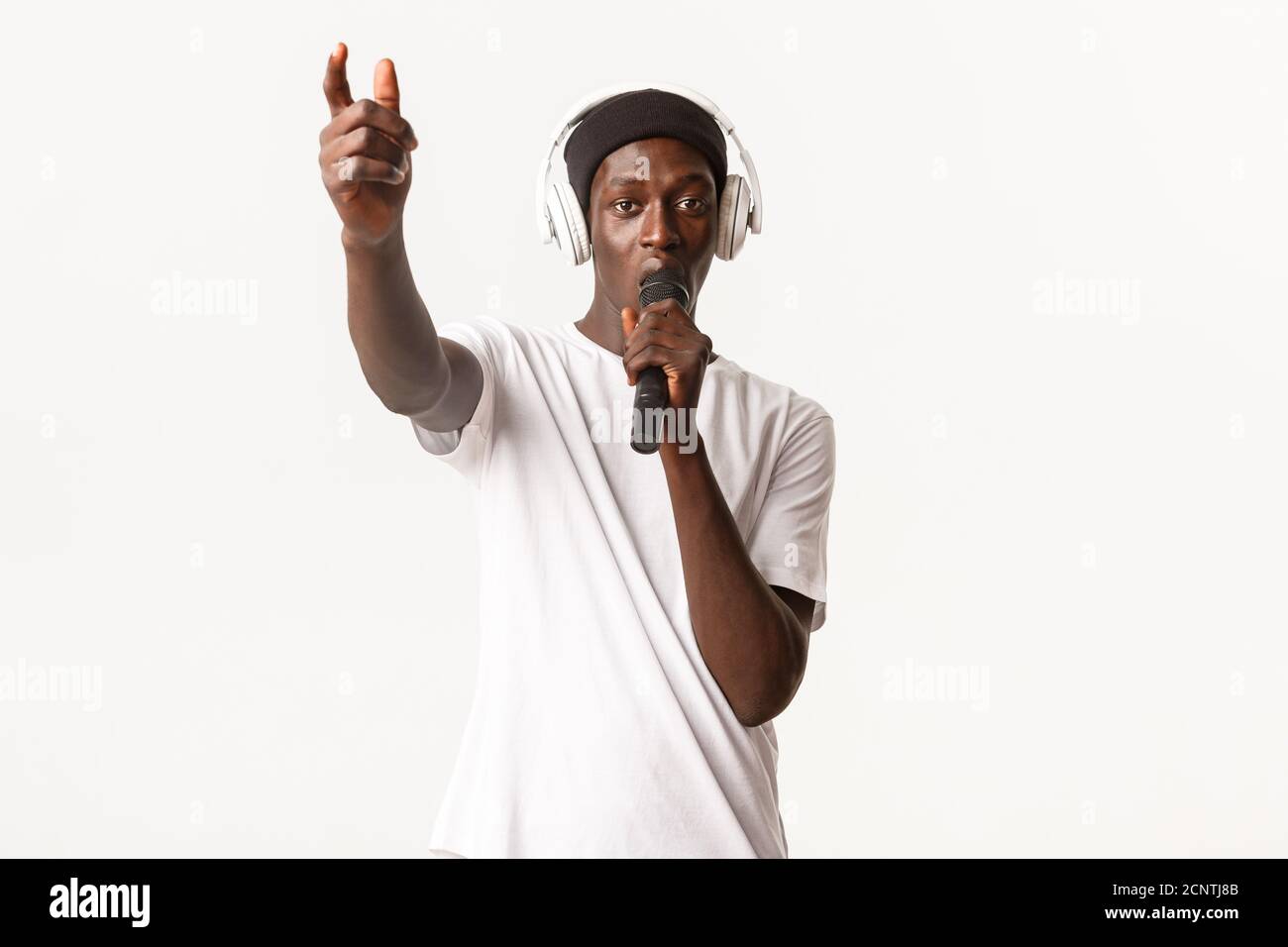 Portrait of sassy and cool african-american guy in wireless headphones, singing into microphone, standing white background Stock Photo