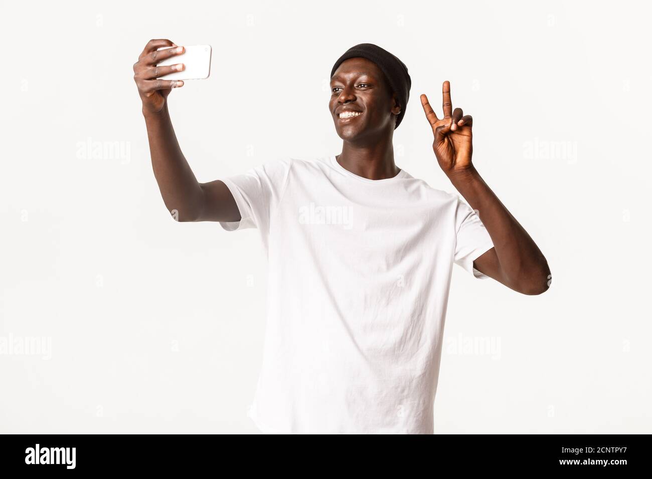 Portrait of cheerful handsome Black in beanie, taking selfie on mobile phone and showing peace gesture happy, smiling over white background Stock Photo - Alamy