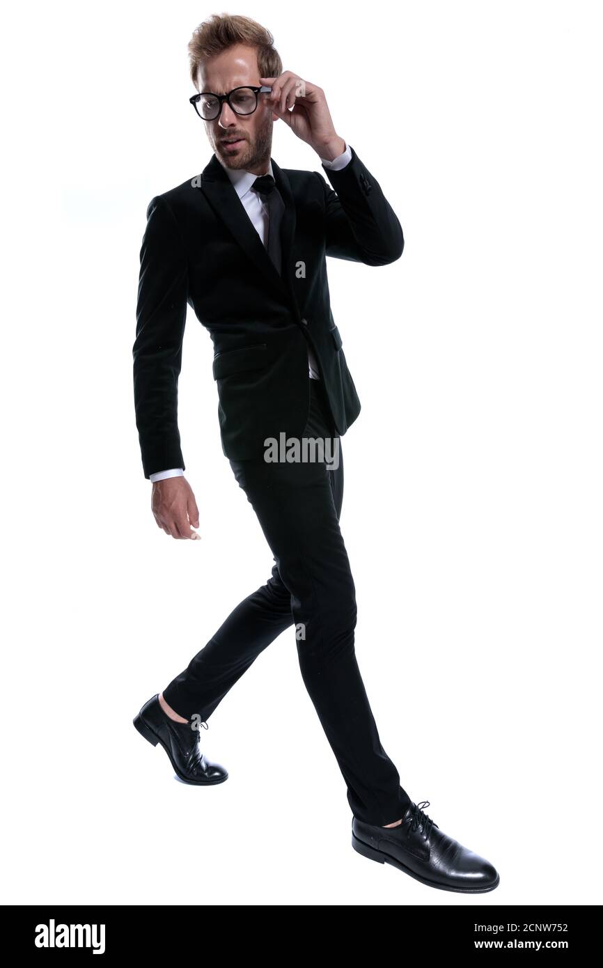 dramatic frowning man in black suit adjusting glasses, looking to side and confidently walking isolated on white background Stock Photo