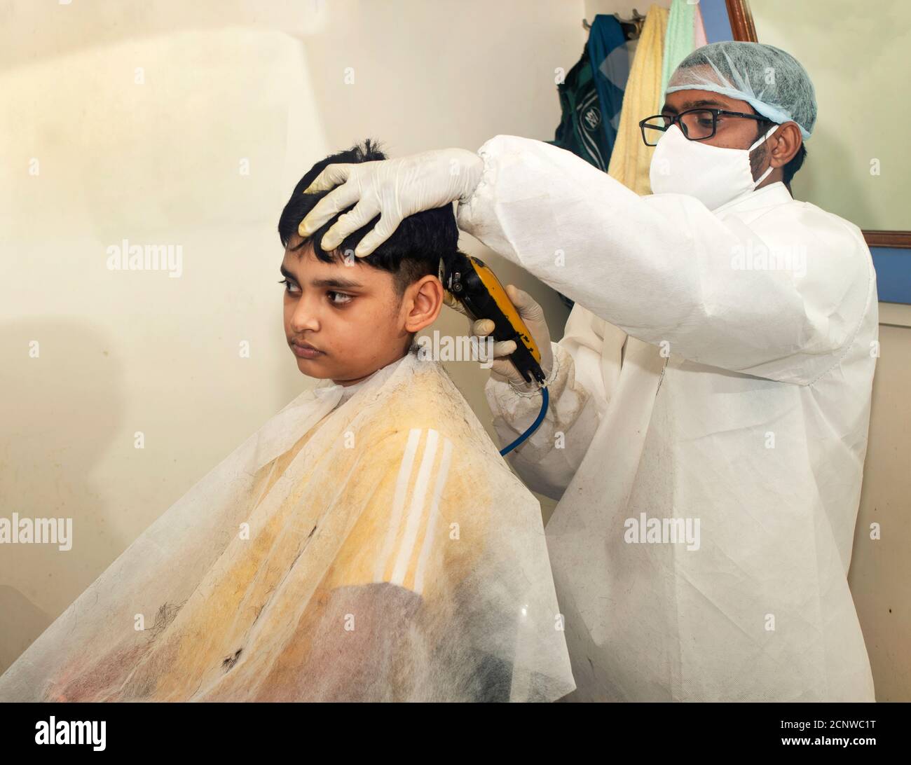 Boy getting a haircut at the barber shop. Barber wearing a white suit and mask and gloves. Location: Nashik, Maharashtra, India Stock Photo