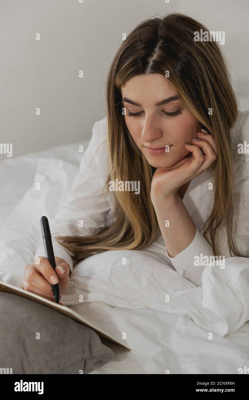 Beautiful young woman laying on bed and writing a diary in bedroom at