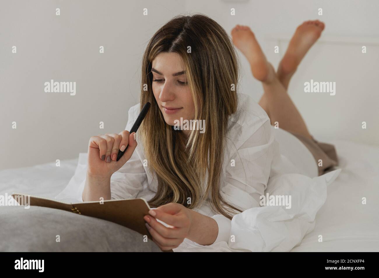 Beautiful young woman laying on bed with black pen and looking on brown notebook in bedroom at home. Lifestyle image of girl thinking with pen and not Stock Photo