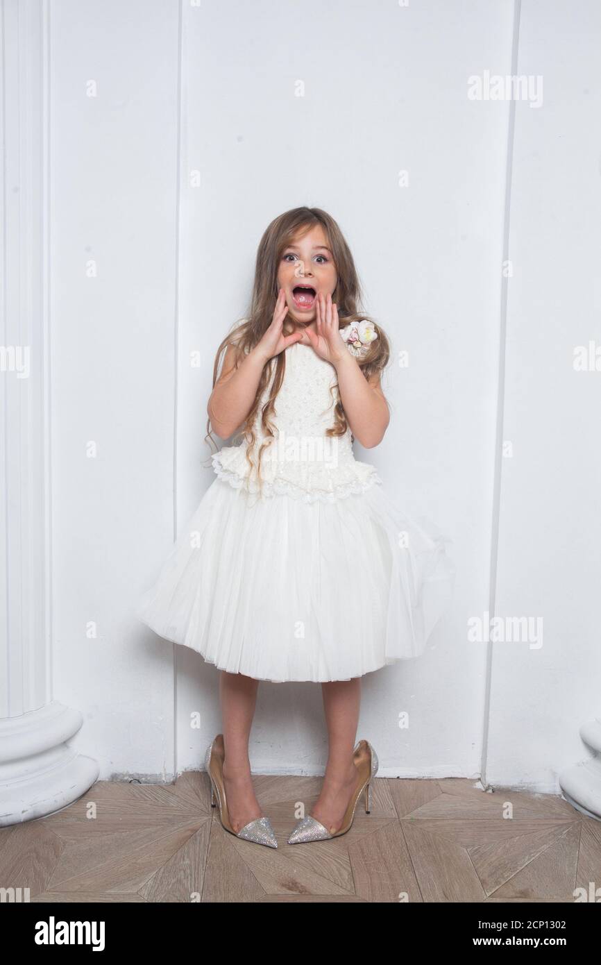 Little princess - excited emotional cute girl in fashion white dress having fun and wearing big mothers sparkle high heels shoes on white background Stock Photo