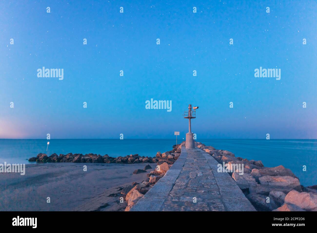 Small lighthouse or navigation beacon on the end of a seawall on Grado island, Italy as a guide to shipping at sunset over a calm sea. Province of Stock Photo