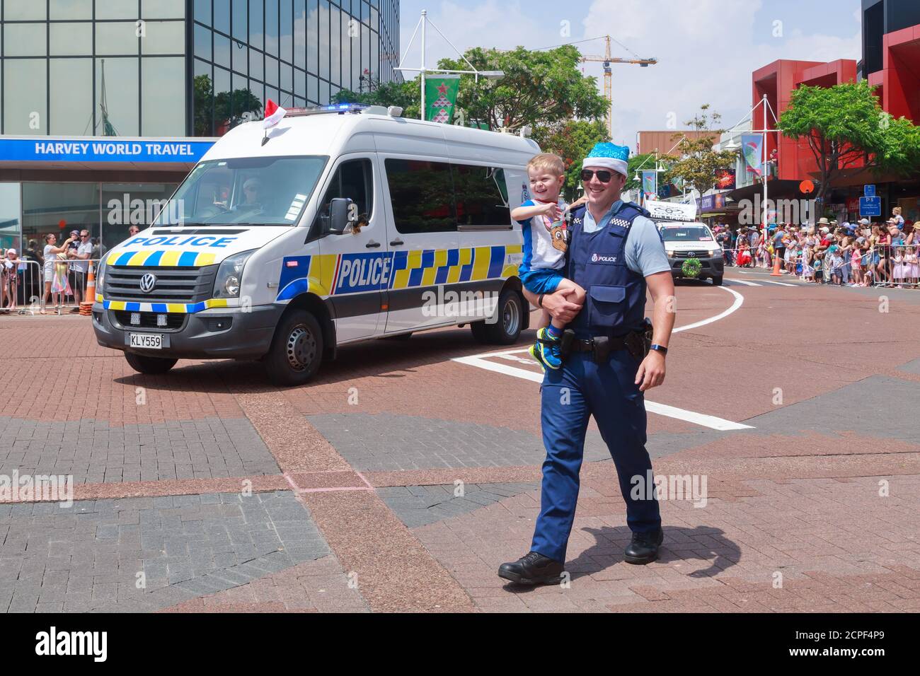 A New Zealand police officer carrying a child at a Christmas parade. Driving beside him is a police van. Tauranga, New Zealand, November 30 2019 Stock Photo