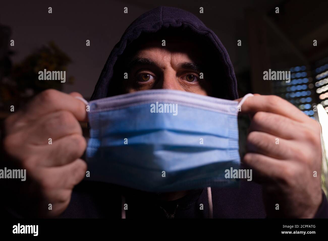 Man in dark hoodie holding blue surgical mask in front of his face ...