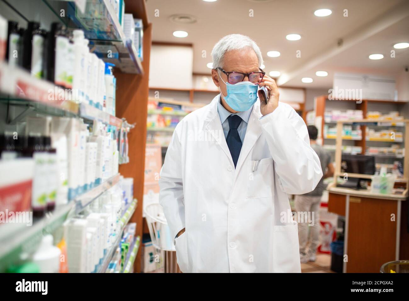 Masked pharmacist talking on the phone while walking in his pharmacy, coronavirus concept Stock Photo