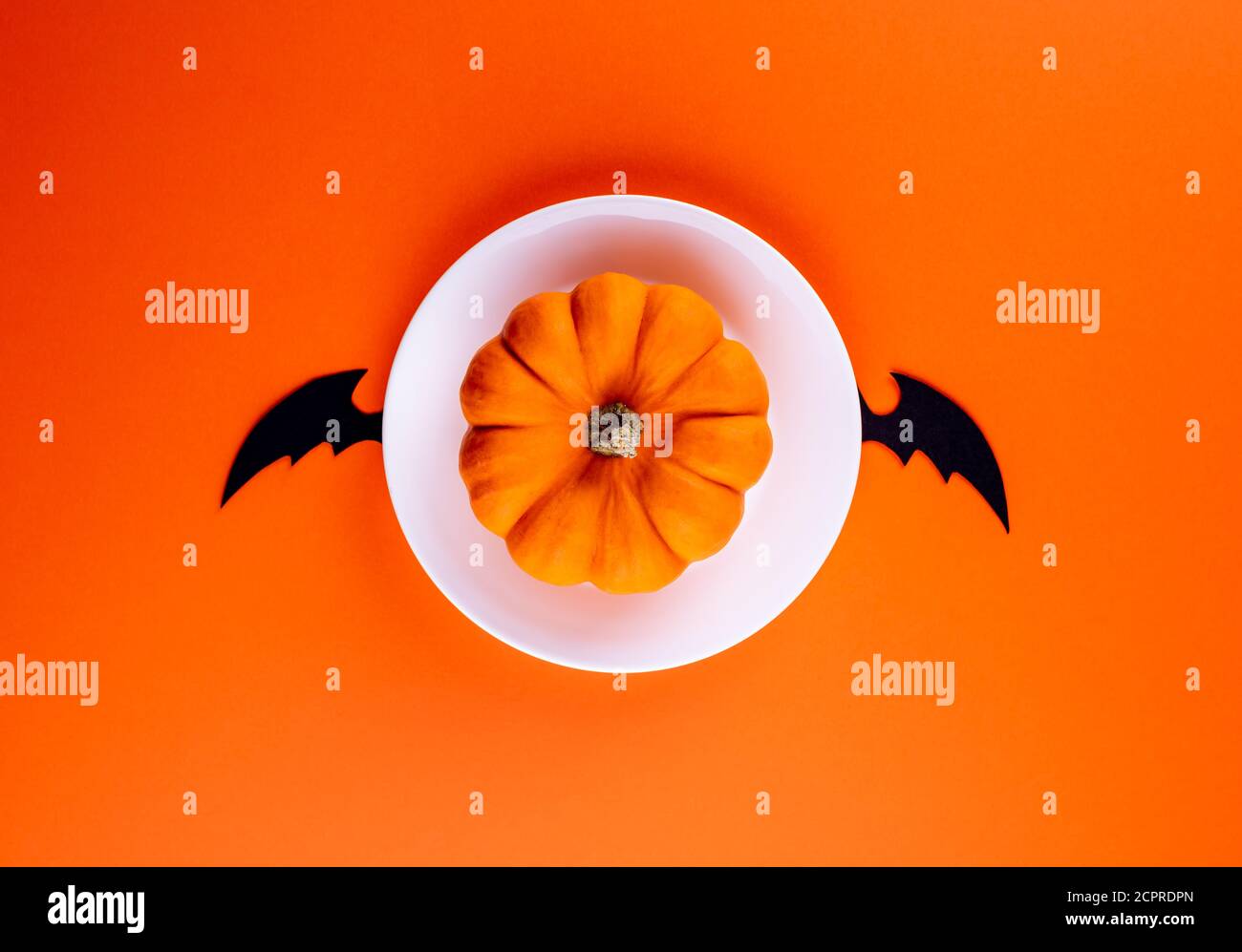 Whole raw orange pumpkin on white round plate with hand carved black bat wings in center of horizontal orange paper background. Copy space. Creative f Stock Photo