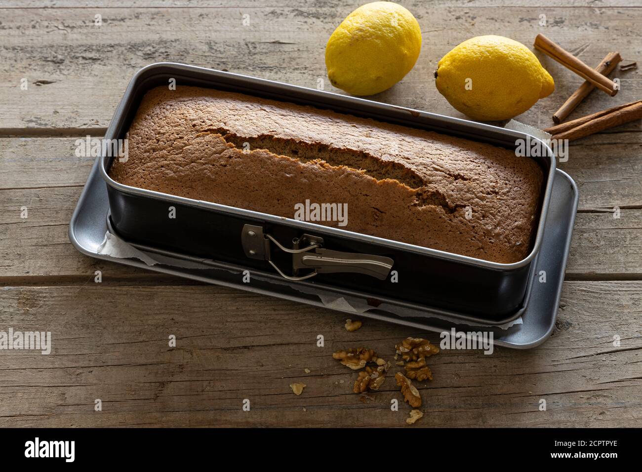 Hot homemade cake straight from the oven in a baking tin surrounded by lemon, cinnamon, and walnuts and placed on a vintage wooden table. Stock Photo