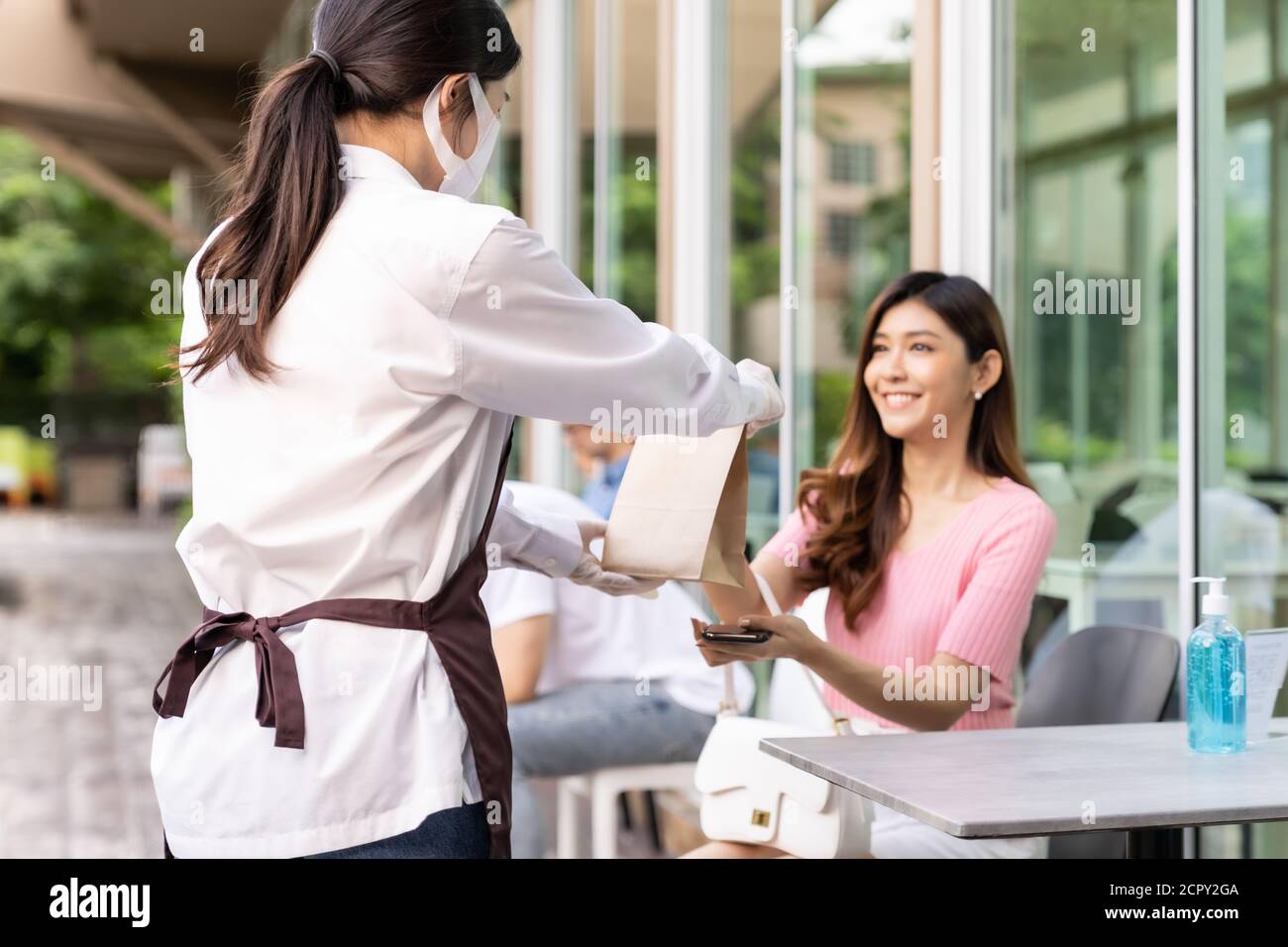 Back view of asian waitress with face mask give order of take out food bag to attractive woman female customer. Take away or take-out food service con Stock Photo