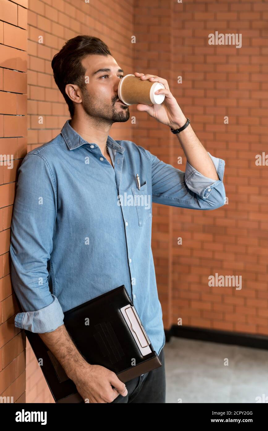 Portrait of middle east caucasian businessman drinking coffee from takeout disposable coffee cup and clipboard in meeting room after lunch break. Stock Photo