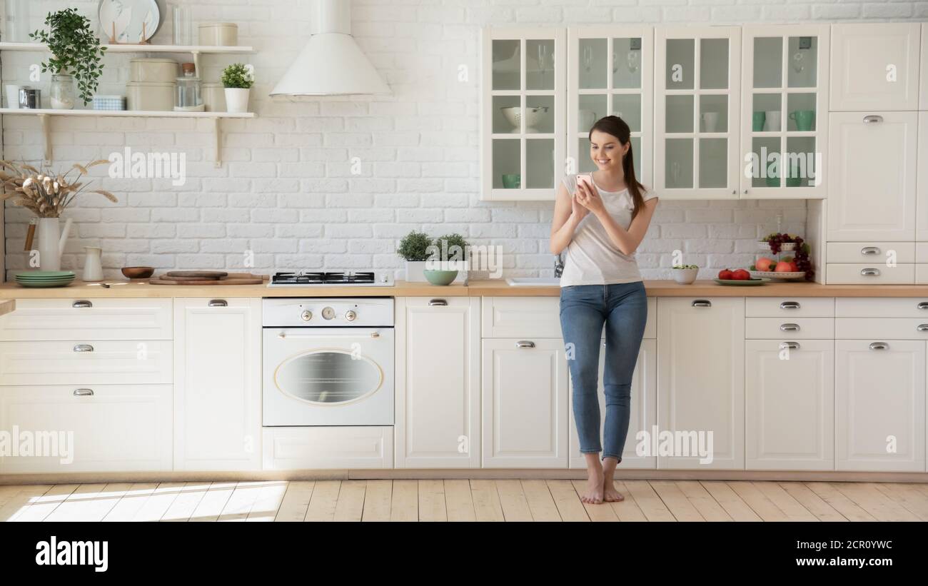 Positive young lady using cell phone in the kitchen Stock Photo