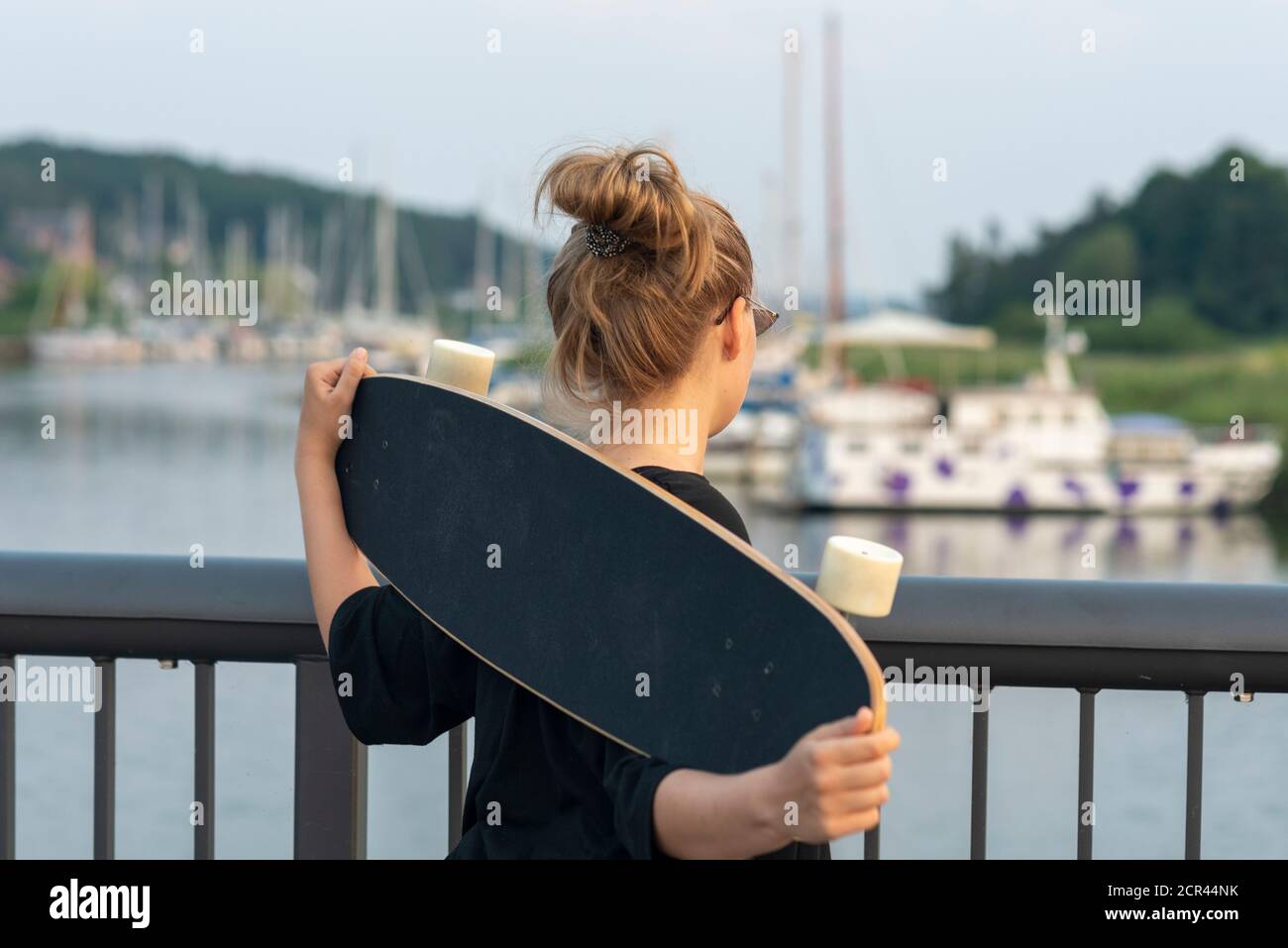 Girl stands with a longboard on a bridge in a sailing harbor Stock Photo