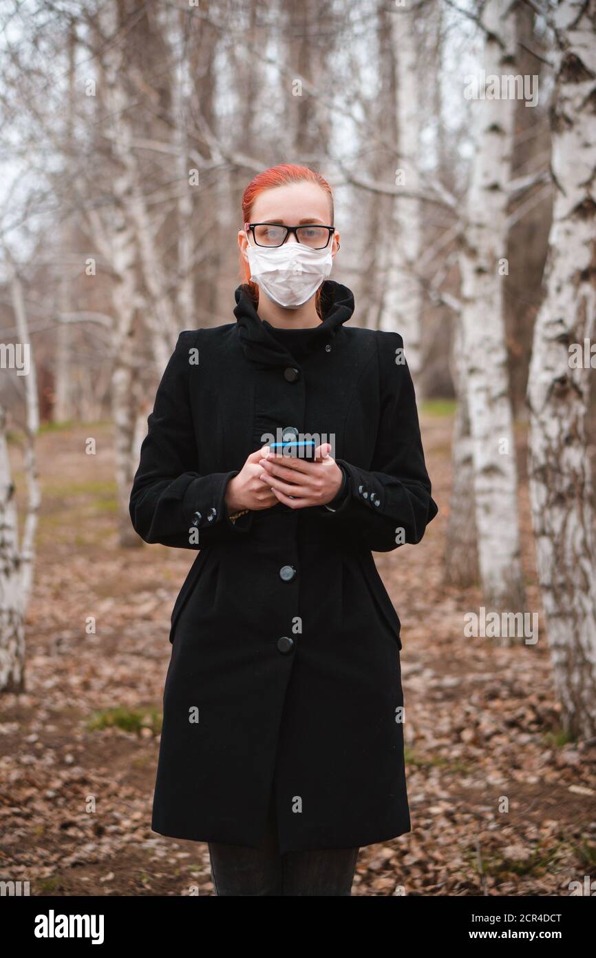 Young beautiful woman in medical mask holds phone in hand in park and looks at camera. Health care or epidemic concept. Stock Photo