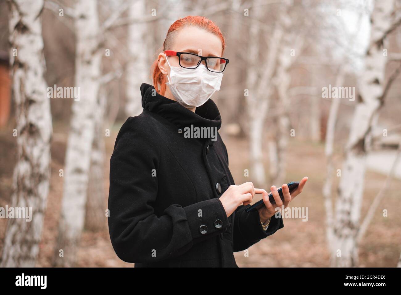 Coronavirus and health care concept with space for text. Young woman in mask holds phone in hand in park and looks at camera. Stock Photo