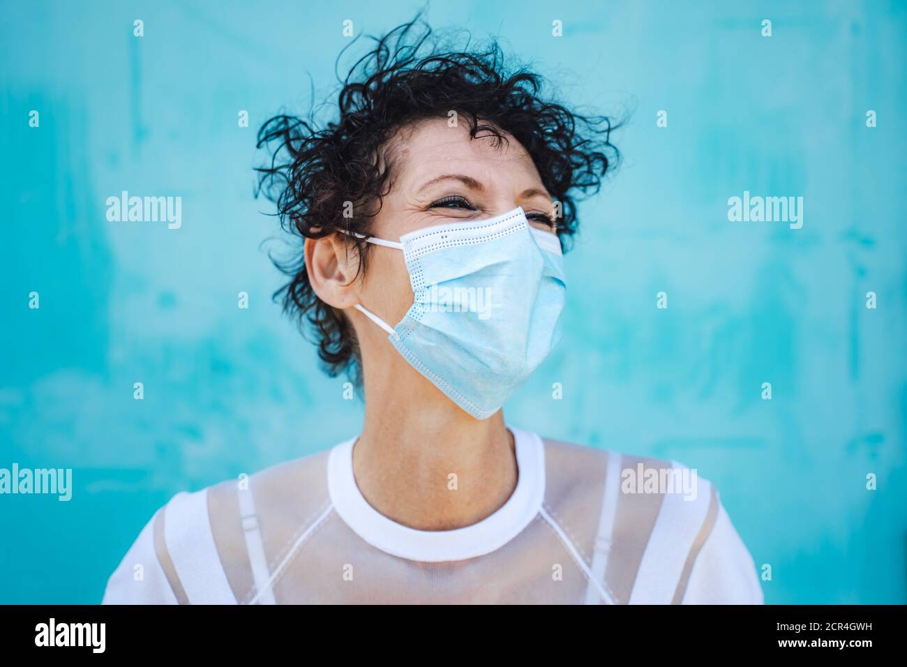 Close up of mature woman with curly short hair wearing safety mask against blue wall Stock Photo