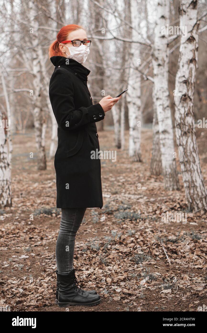 Young beautiful woman in medical mask holds phone in hand in park and looks at camera. Health care or epidemic concept. Stock Photo