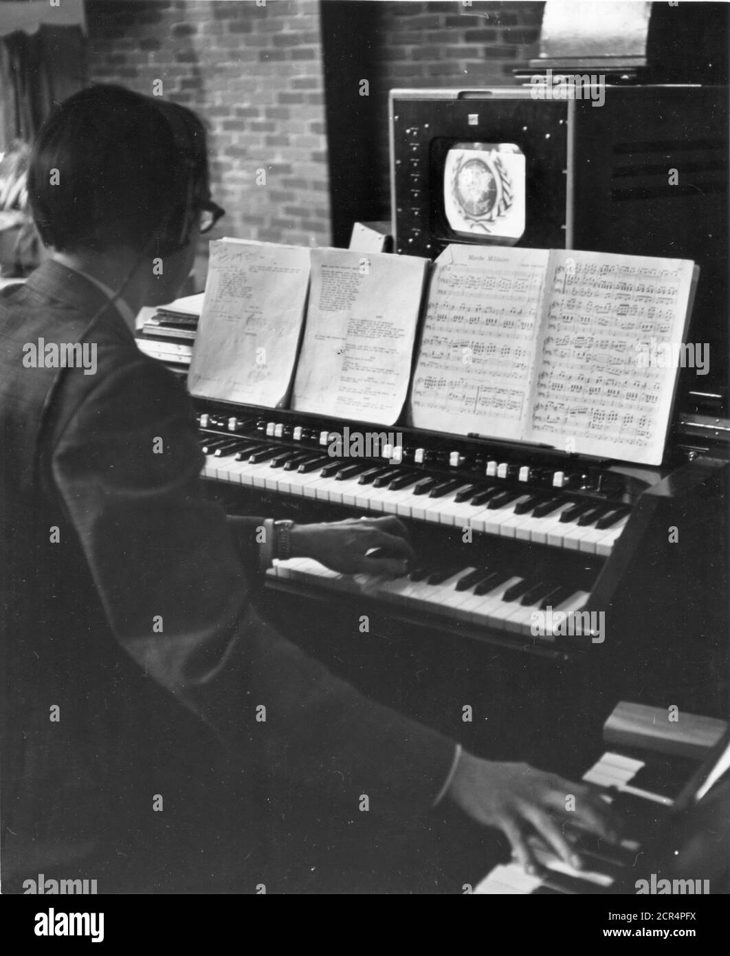 The organist providing background music for a television show watches the image on the monitor above the organ keyboard, listens to cues from the control room through earphones, has both a script and a musical score to watch and both hands full operating two keyboards, New York, NY, 7/1/1949. (Photo by RBM Vintage Images) Stock Photo