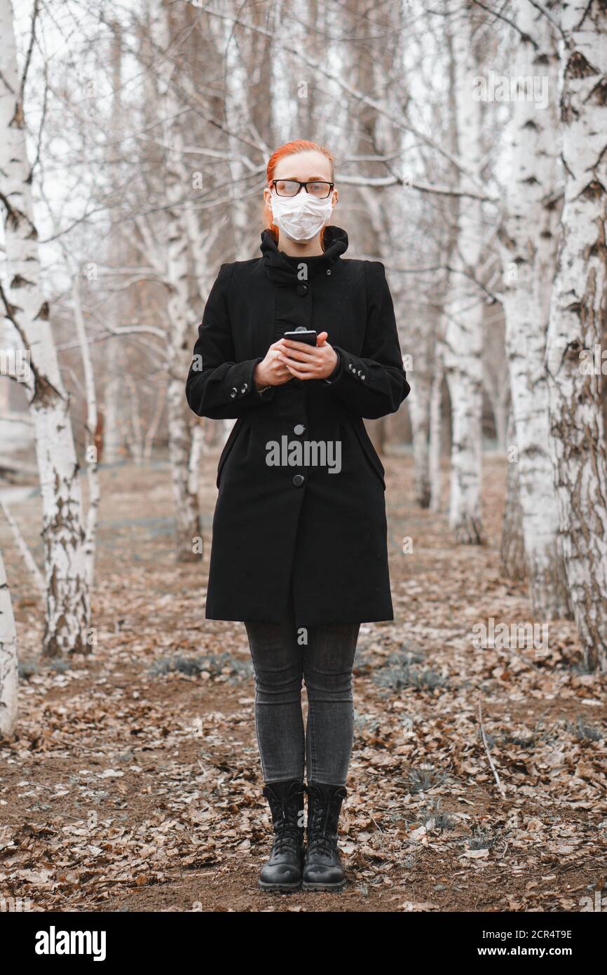 Girl in medical mask with phone in her hand stands on street and looks at camera. Virus protection concept. Stock Photo