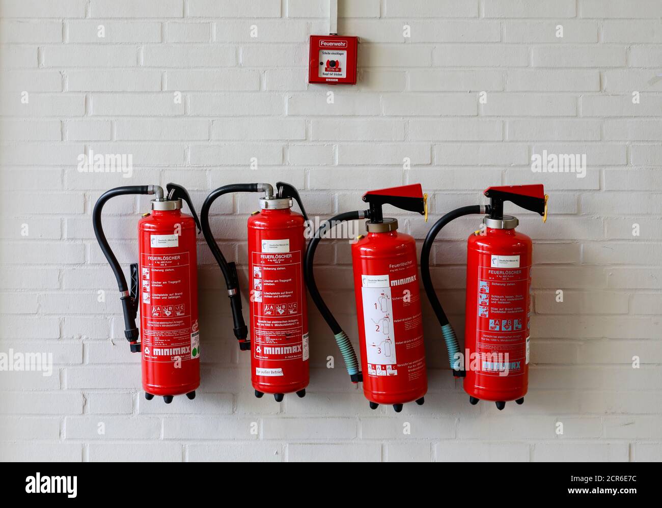 Hannover, Lower Saxony, Germany - Four red fire extinguishers on a wall under a fire alarm at the Hannover Messe. Stock Photo